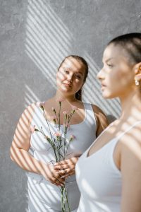 2 women standing in the diffused light of window and blinds, one holding a stems of flowers.